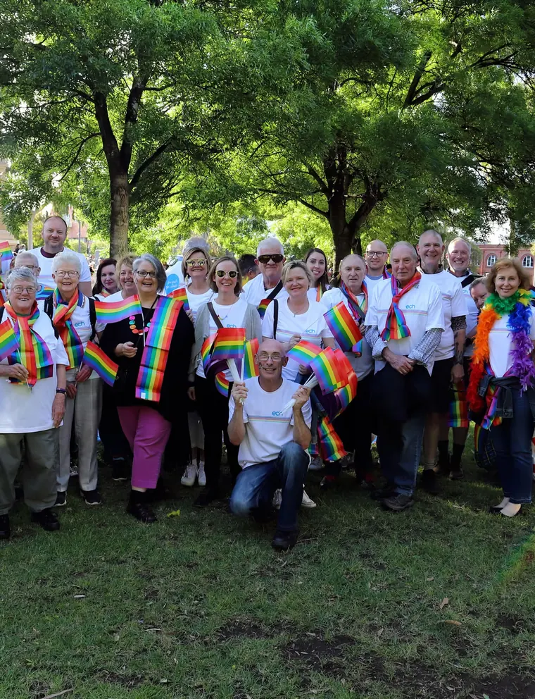 Community members holding rainbow flags at an outdoor gathering under trees.